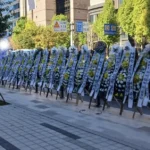 Funeral flowers have become a symbol of protests among Korean fans, used to express strong disapproval. Recent incidents involved protests against Seunghan’s return to RIIZE and Suga’s DUI case, with fans sending funeral wreaths to demand their removal.